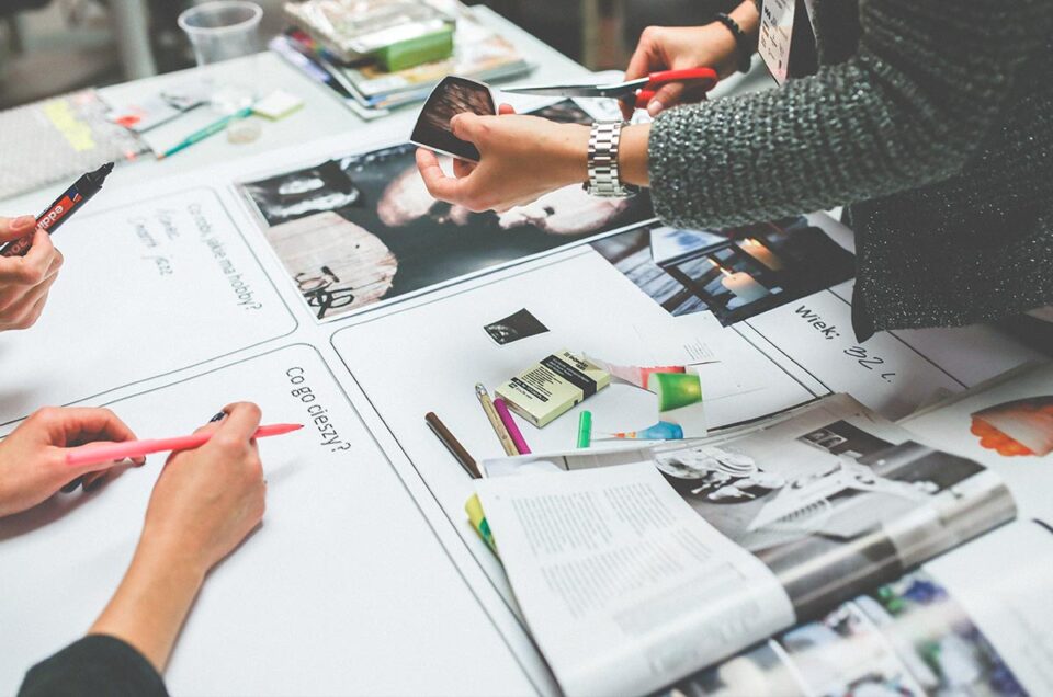 Zdjęcie przedstawia rozłożone gazety na których pracują trzy osoby Widzimy trzy pary rąk które trzymają długopisy