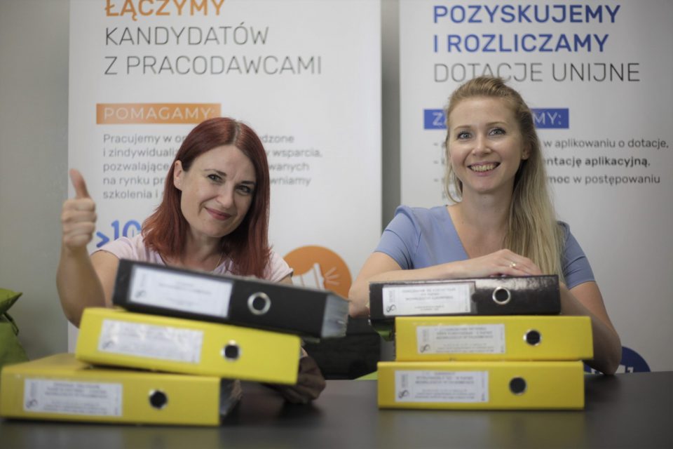 dwie kobiety siedzą za segregatorami mają uśmiechnięte buzie a w tle banery reklamowe