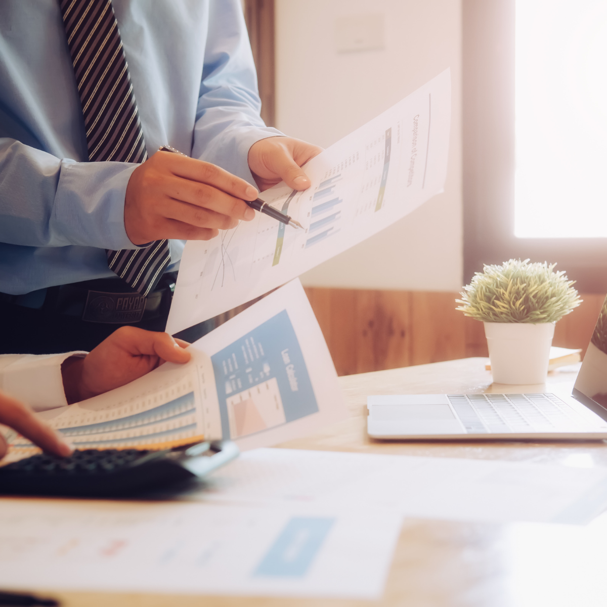 mężczyzna pokazuje pewien wykres partnerowi w pracy a na stole leży laptop i roślinka