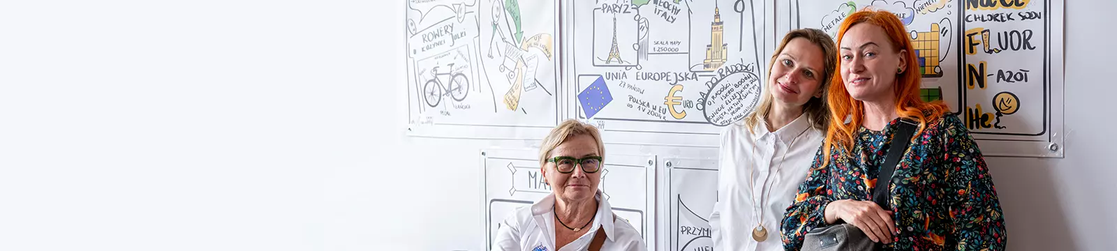 trzy kobiety w tym jedna siedzi a z tyłu ściana z motywami europejskimi
