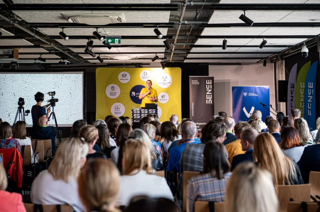 mężczyzna w żółtej koszulce prowadzi prezentację mówi do mikrofonu ludzie siedzą na krzesłach i go słuchają