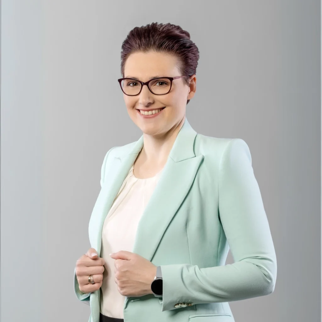Kobieta w okularach i miętowej marynarce, z krótkimi ciemnymi włosami, pozuje do zdjęcia z uśmiechem.