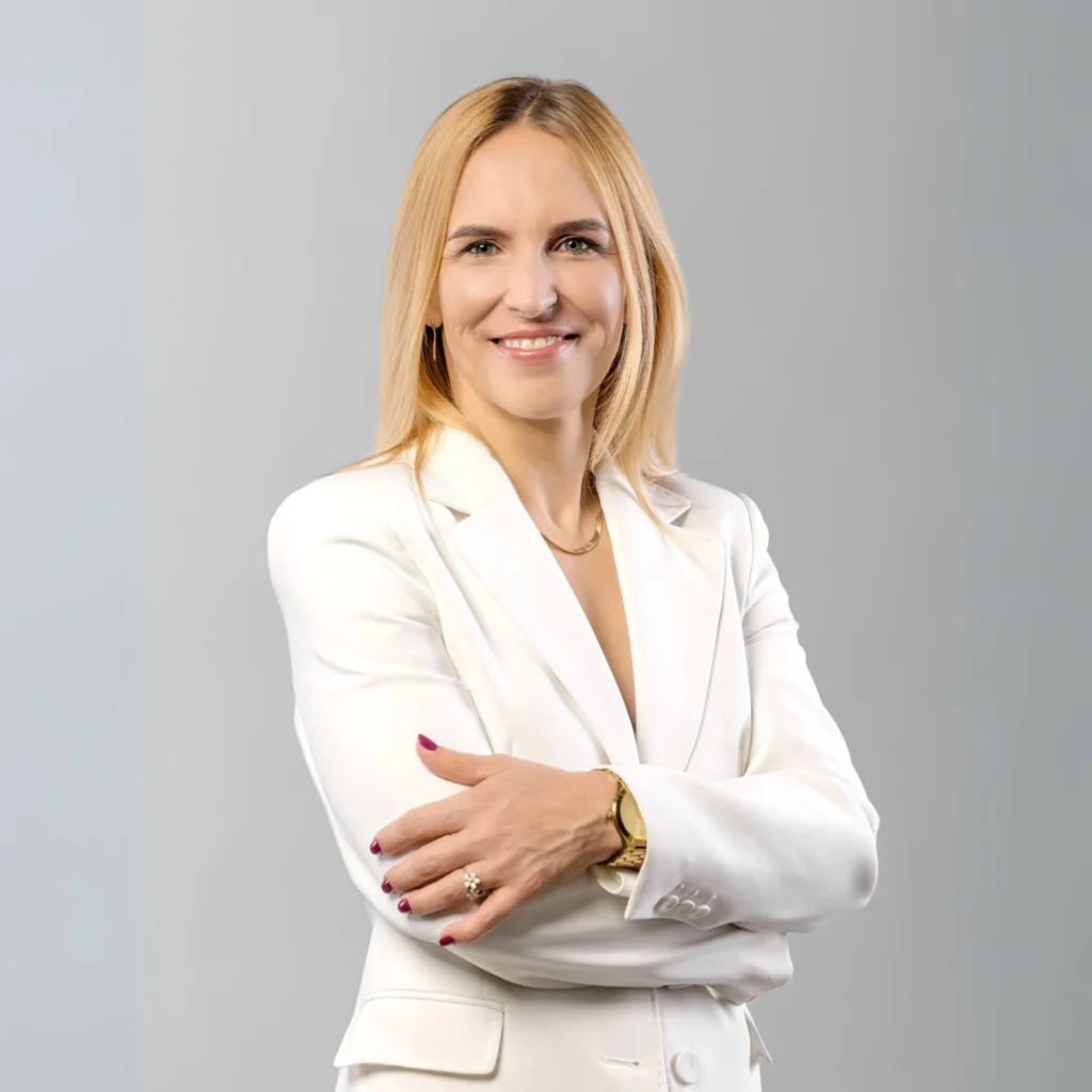 Elegancka kobieta z długimi blond włosami w białym garniturze, stoi z założonymi rękoma i uśmiecha się.