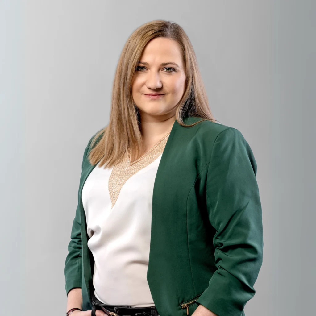 Uśmiechnięta kobieta w zielonym żakiecie i białej koszuli, stojąca na neutralnym tle.
