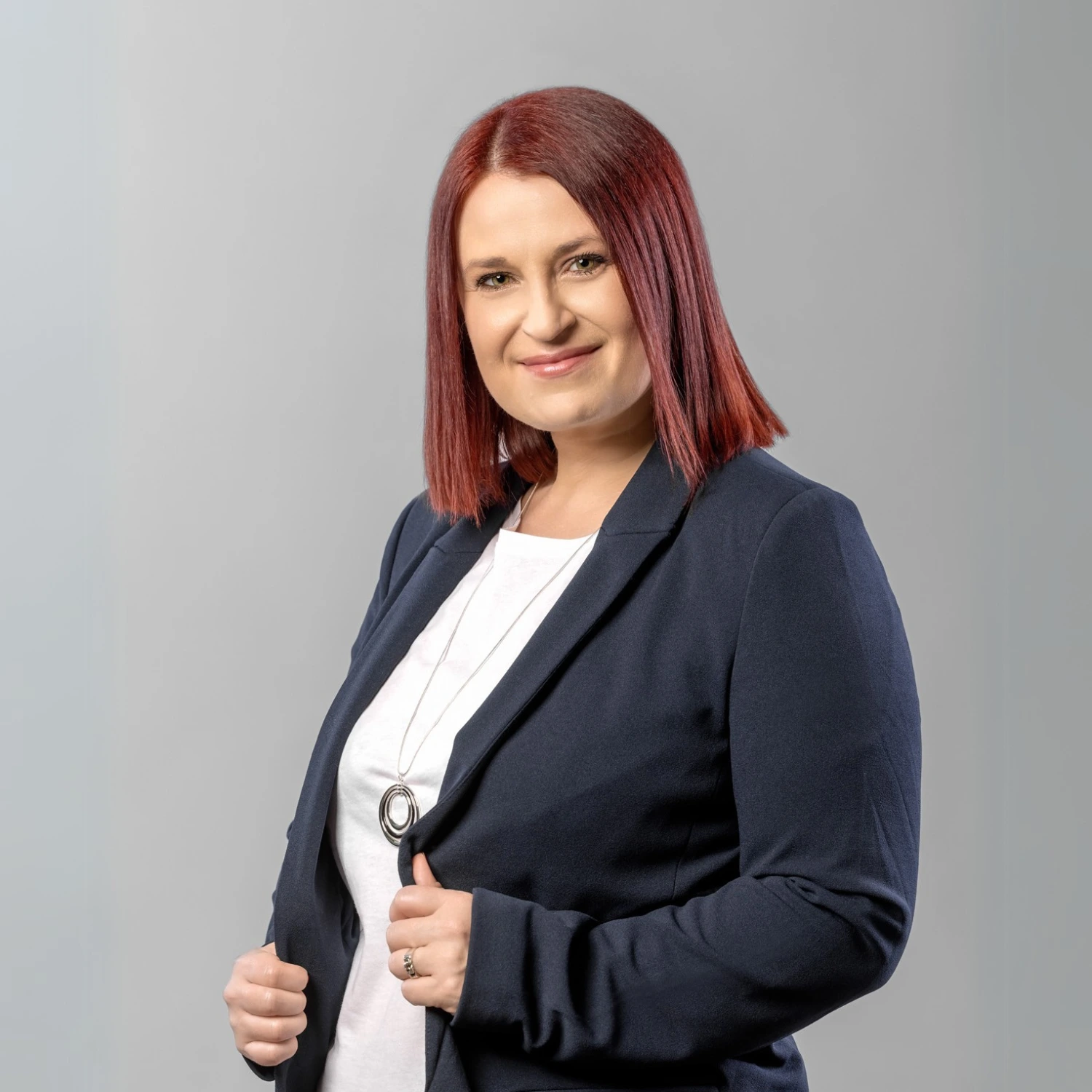 Anna Wiśniewska Kobieta z czerwonymi włosami, ubrana w białą koszulę, stoi na tle neutralnego tła i lekko się uśmiecha.