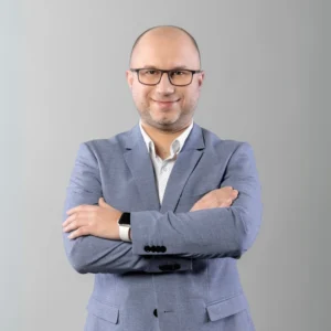 Łysy mężczyzna w okularach i szarej marynarce stoi z rękoma założonymi na piersi i uśmiecha się.