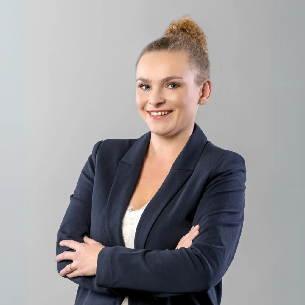 Kobieta w koku i ciemnej marynarce pozuje do zdjęcia, uśmiechając się i prezentując profesjonalny wizerunek.