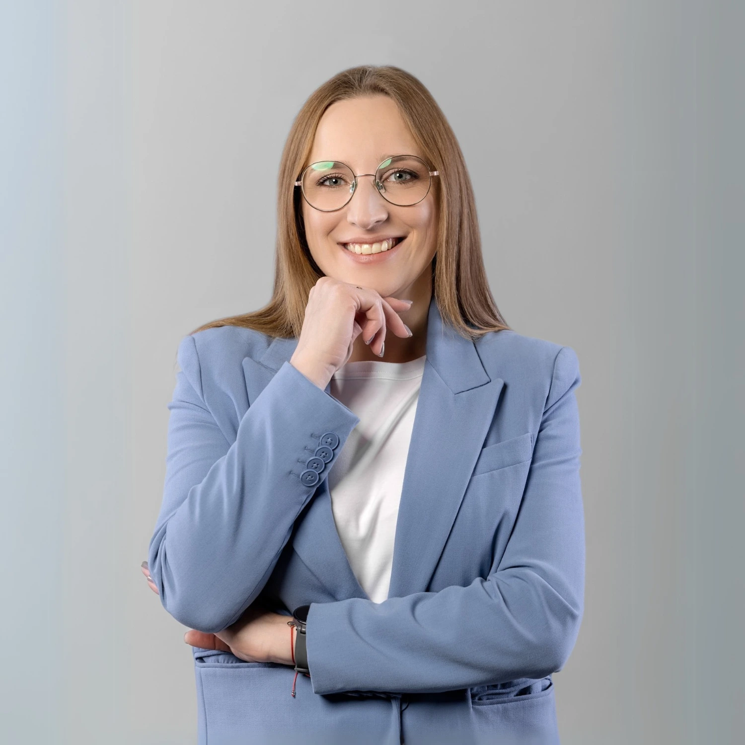 Kobieta w niebieskiej marynarce i okularach, uśmiecha się do obiektywu, podpierając podbródek prawą dłonią.