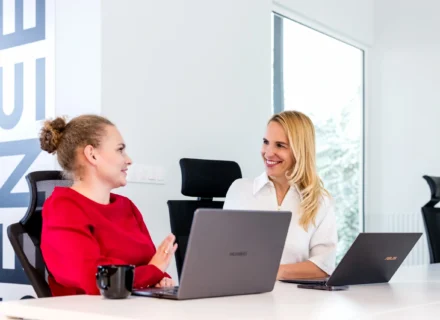 dwie kobiety siedzą przy biurku z laptopami rozmawiają