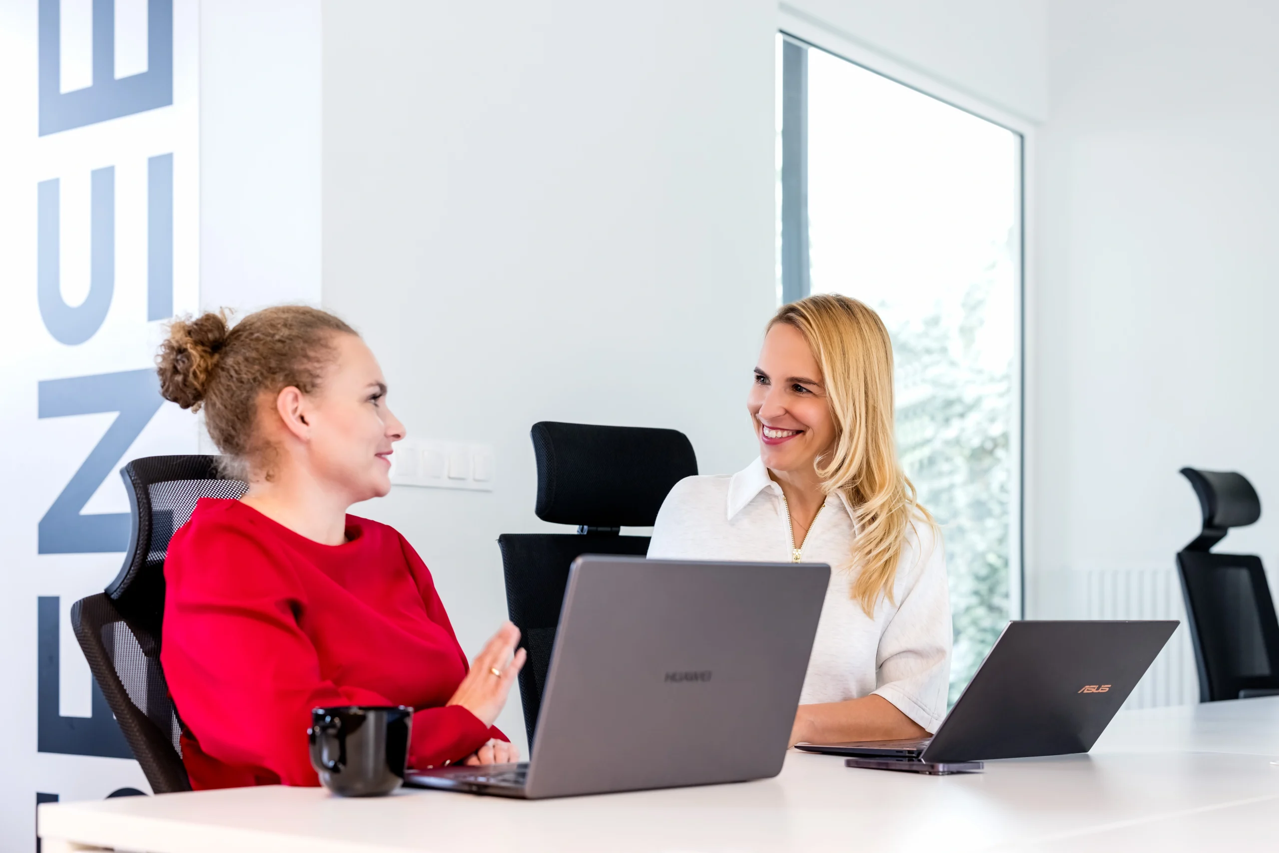 dwie kobiety siedzą przy biurku z laptopami rozmawiają