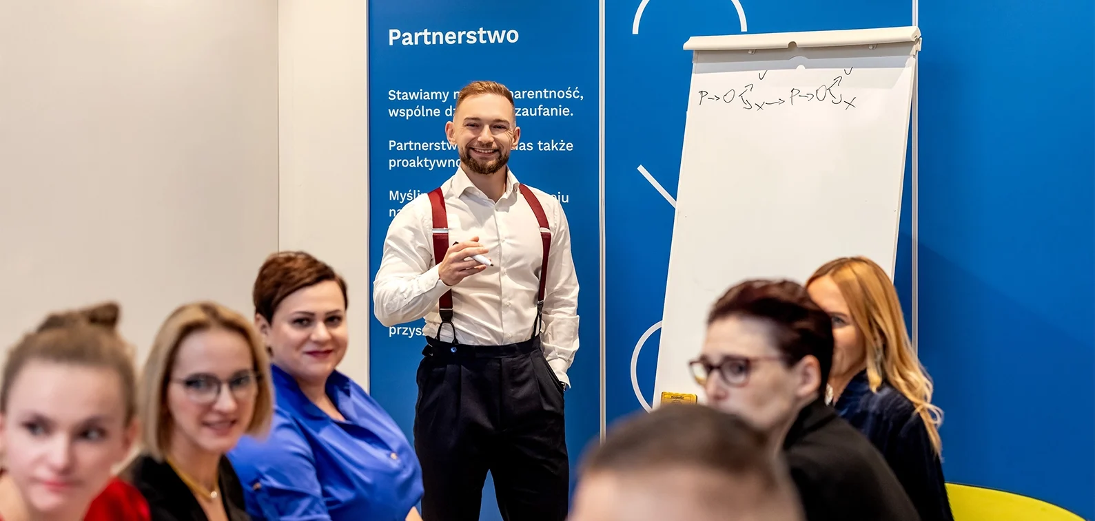 Mężczyzna w białej koszuli stojący na przed niebieską ścianą przed grupą osób n