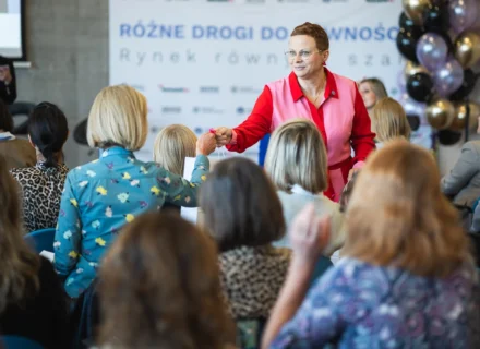 Kobieta w różowo czerwonym stroju wręcza upominek uczestniczce konferencji na tle ścianki z napisem Różne drogi do równości