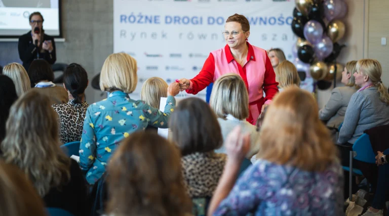 Kobieta w różowo czerwonym stroju wręcza upominek uczestniczce konferencji na tle ścianki z napisem Różne drogi do równości
