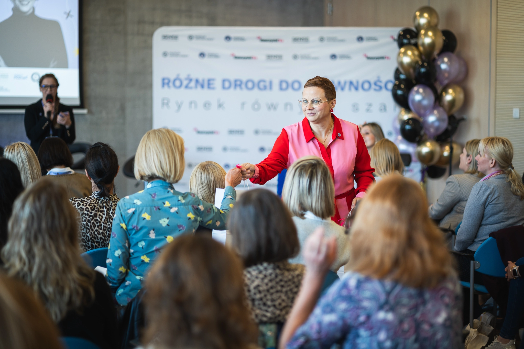 Kobieta w różowo czerwonym stroju wręcza upominek uczestniczce konferencji na tle ścianki z napisem Różne drogi do równości