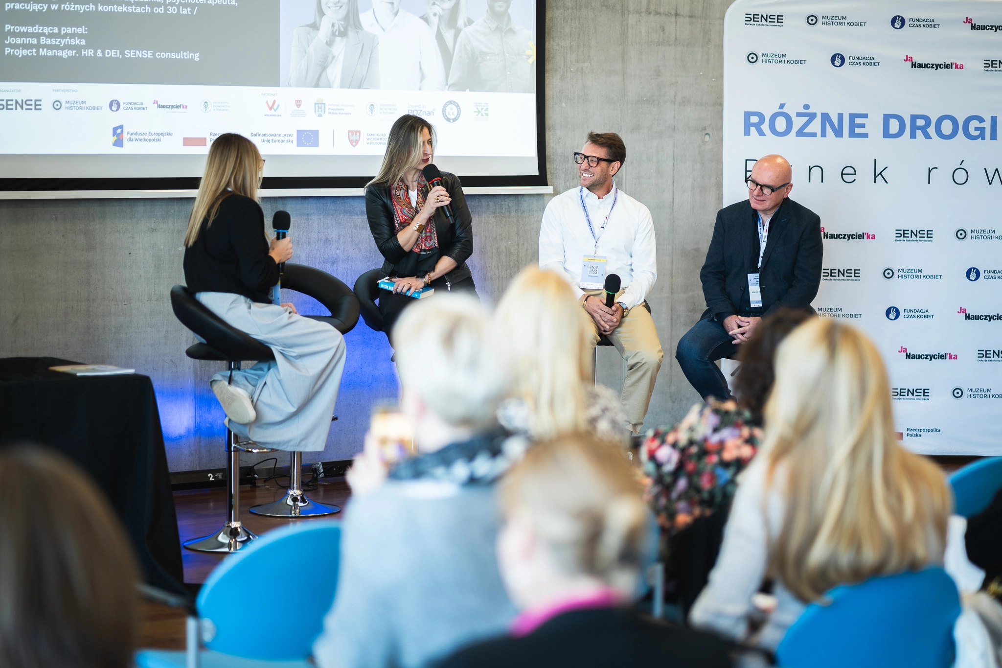 Panel dyskusyjny z udziałem czterech osób na scenie siedzących na hokerach na tle ekranu z prezentacją i banerów z logami organizatorów w tym SENSE