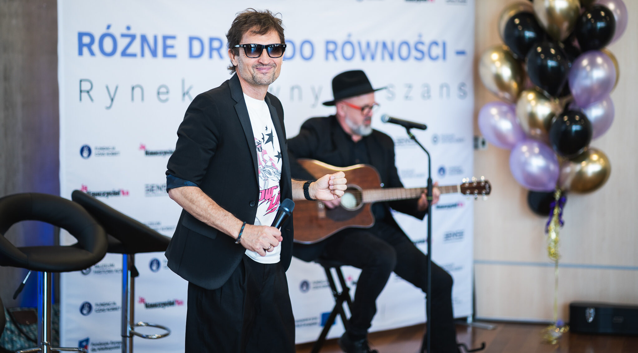 Mężczyzna w okularach przeciwsłonecznych czarnej marynarce i białym tiszercie trzyma mikrofon i uśmiecha się na tle ścianki z napisem Różne drogi do równości Rynek pełen szans oraz logo Grupa SENSE