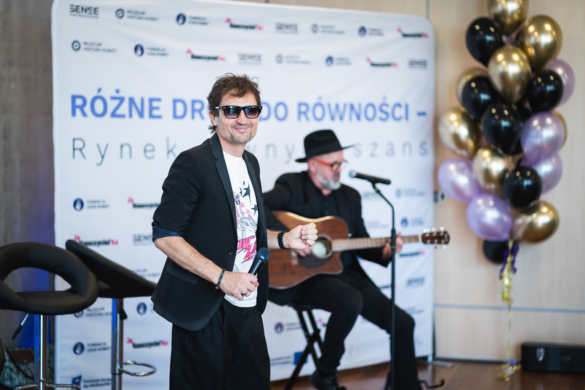 Mężczyzna w okularach przeciwsłonecznych czarnej marynarce i białym tiszercie trzyma mikrofon i uśmiecha się na tle ścianki z napisem Różne drogi do równości Rynek pełen szans oraz logo Grupa SENSE