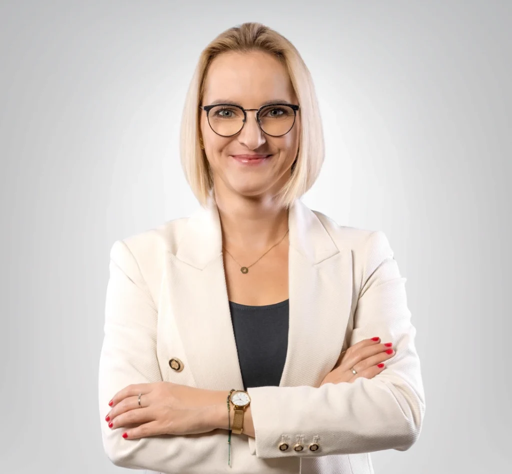 Portret kobiety w beżowej marynarce z krótkimi blond włosami i uśmiechem na twarzy