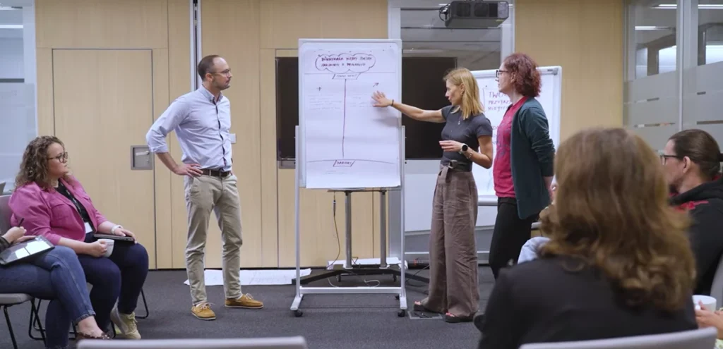 osoby stojące przy tablicy w sali jedna z nich z podniesioną ręką wskazuje na tablice