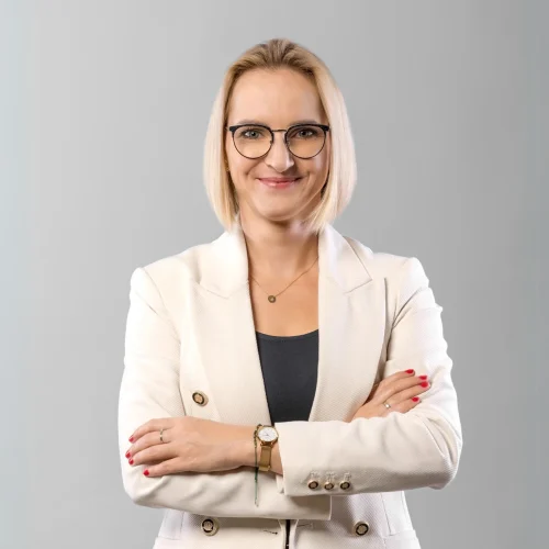 Portret bizneswoman w okularach i jasnej marynarce na szarym tle.