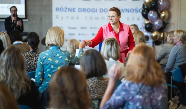 Kobieta w różowo czerwonym stroju wręcza upominek uczestniczce konferencji na tle ścianki z napisem Różne drogi do równości